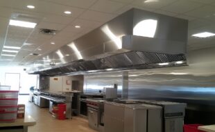 Sistema de exaustor para cozinha industrial em aço inox, instalado sobre equipamentos de cocção em ambiente profissional.