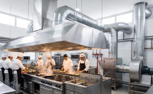 Cozinha profissional com chefs trabalhando sob sistema de coifa e dutos de exaustão para cozinha industrial.
